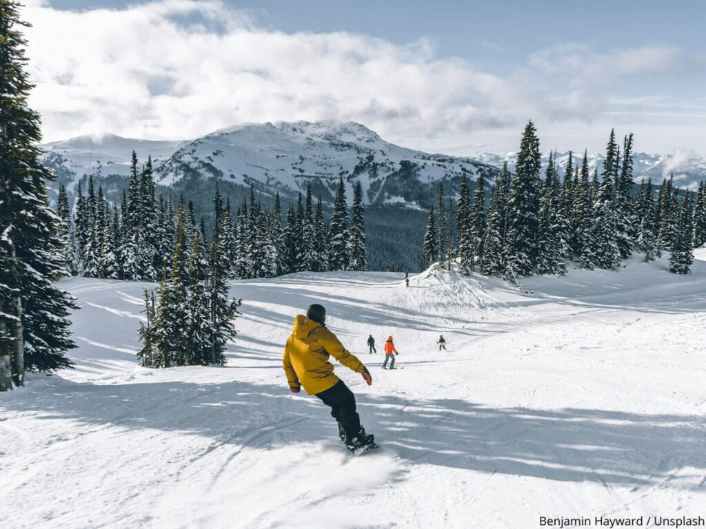 Snowboarder in yellow jacket on a snow slope lined with fir trees and mountains in the distance