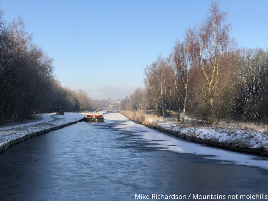 A narrow boat making its way up the Bridgewater Canal with blue skies and ice on the ground